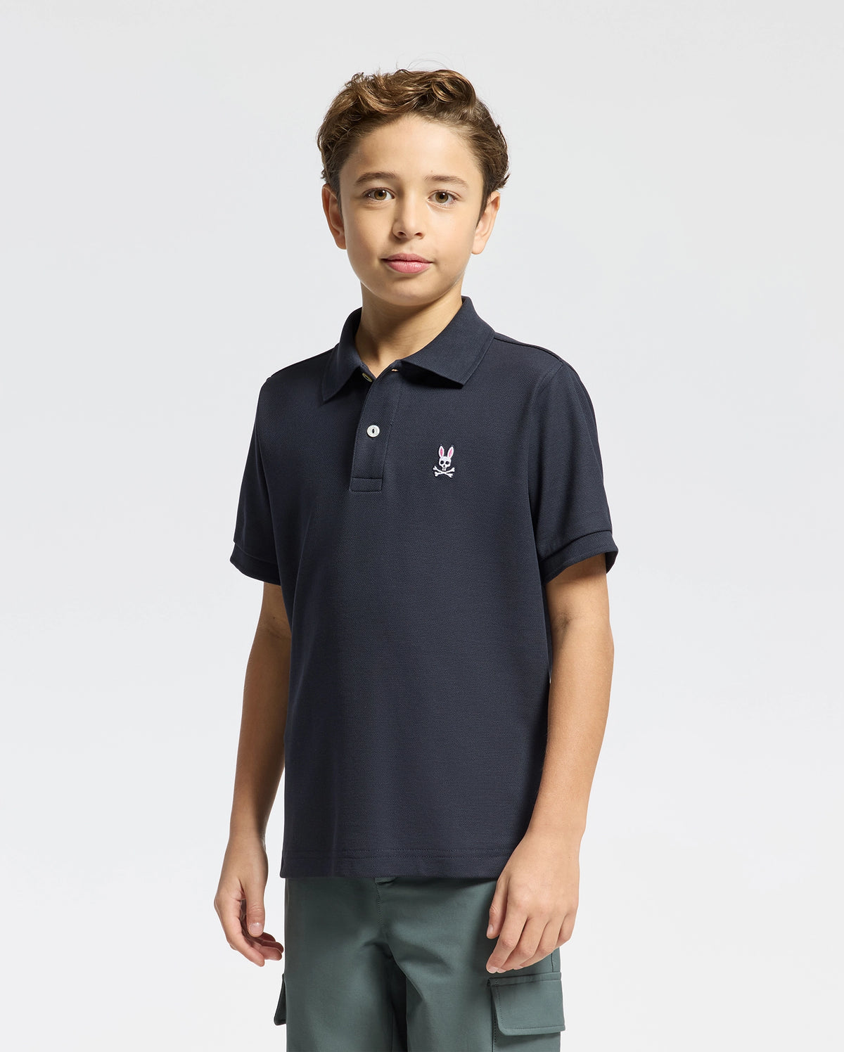 A young boy with short brown hair wears a Psycho Bunny KIDS CLASSIC PIQUE POLO SHIRT (B0K001CRPC) in navy blue Pima cotton with a white chest logo and green cargo shorts, standing against a plain light background.