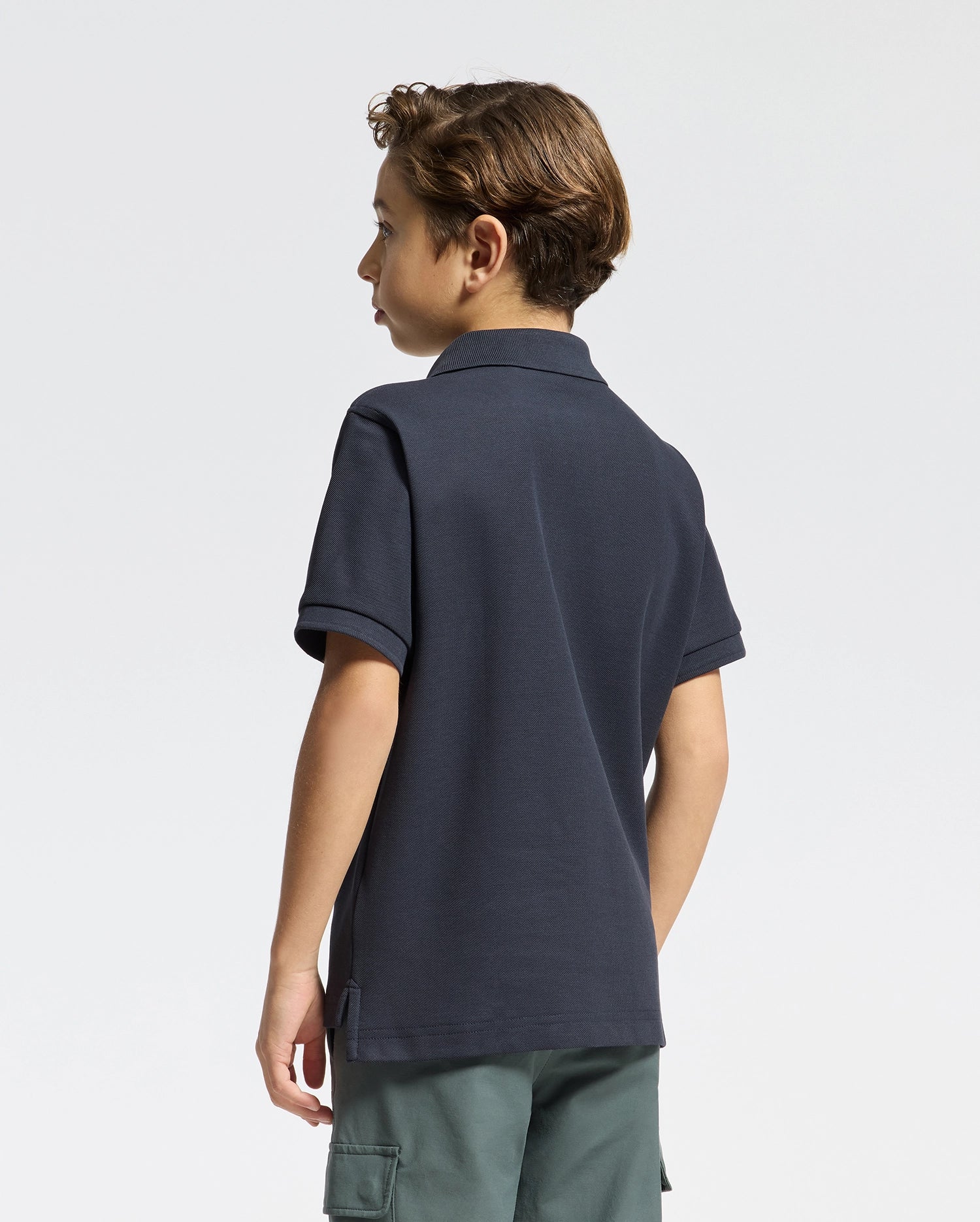 A young boy with short brown hair, seen from behind, is wearing a Psycho Bunny KIDS CLASSIC PIQUE POLO SHIRT - B0K001CRPC in dark navy and green cargo shorts, set against a plain light background.