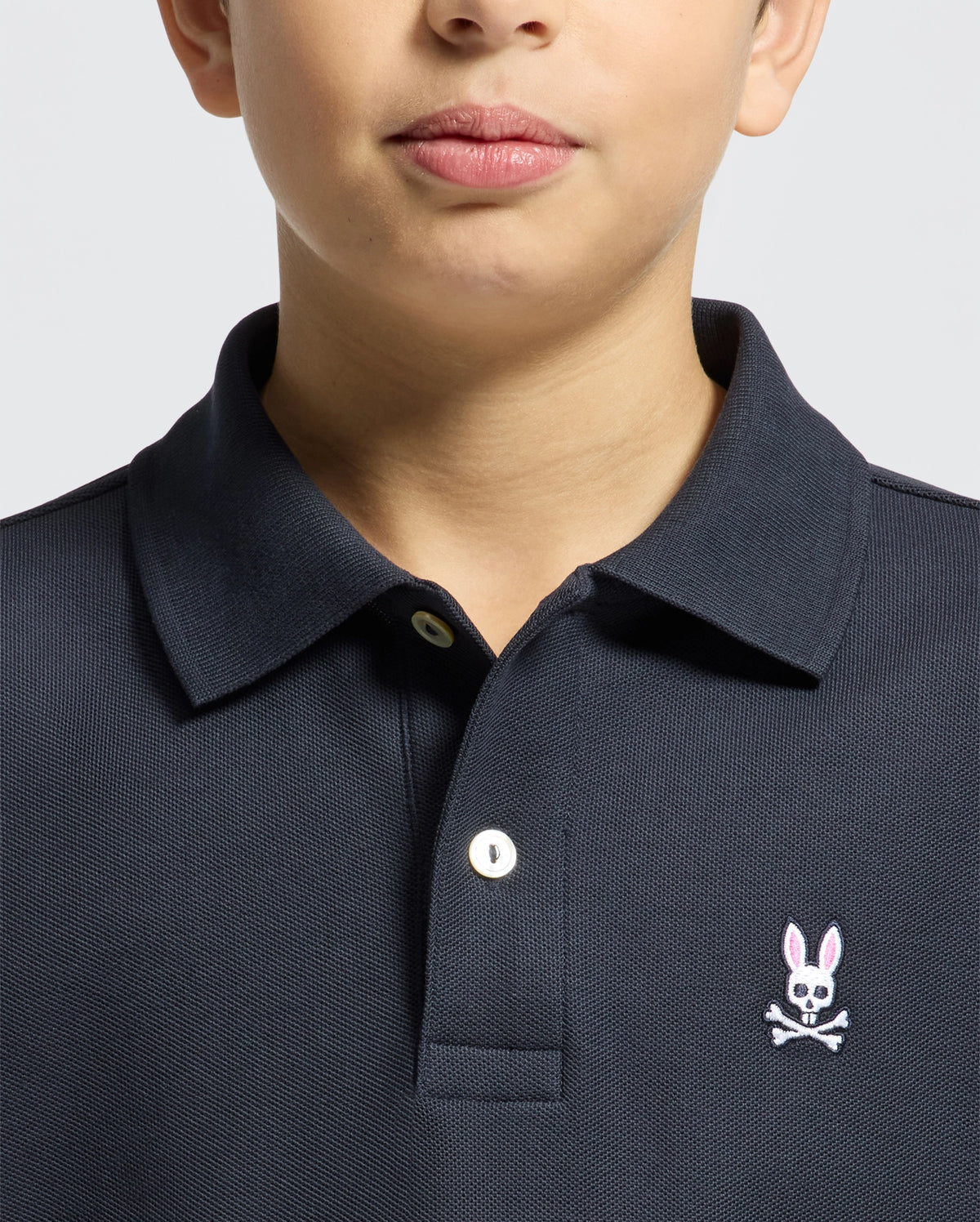 A child wears a Psycho Bunny KIDS CLASSIC PIQUE POLO SHIRT (B0K001CRPC) in dark Pima cotton, with a white button and pink embroidered bunny with crossbones on the left chest; only the lower face and upper torso are shown.
