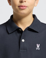 A child wears a Psycho Bunny KIDS CLASSIC PIQUE POLO SHIRT (B0K001CRPC) in dark Pima cotton, with a white button and pink embroidered bunny with crossbones on the left chest; only the lower face and upper torso are shown.