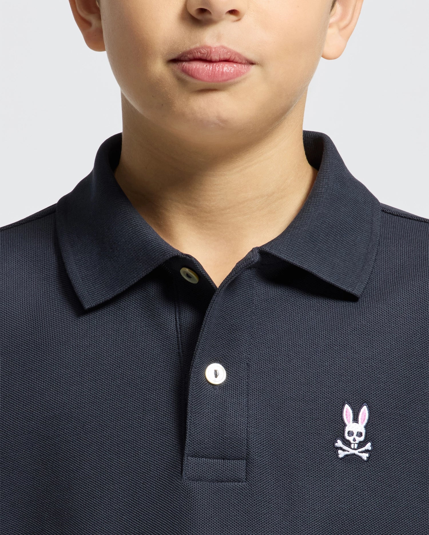 A child wears a Psycho Bunny KIDS CLASSIC PIQUE POLO SHIRT (B0K001CRPC) in dark Pima cotton, with a white button and pink embroidered bunny with crossbones on the left chest; only the lower face and upper torso are shown.