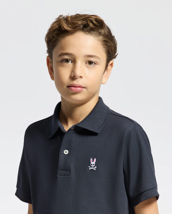 A young boy with light brown hair wears the Psycho Bunny KIDS CLASSIC PIQUE POLO SHIRT - B0K001CRPC in dark navy Pima cotton, featuring a pink bunny and crossbones logo. He stands against a plain light background, looking at the camera neutrally.