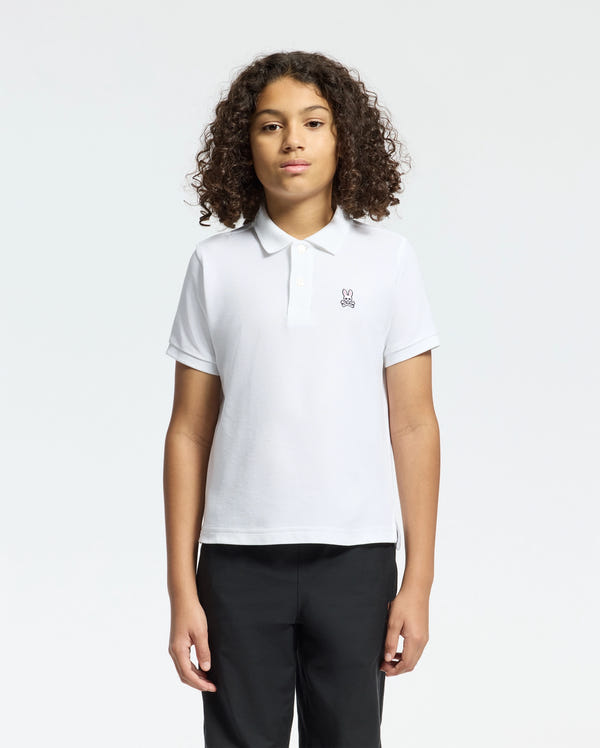 A curly-haired person wearing a Psycho Bunny KIDS CLASSIC PIQUE POLO SHIRT in white Pima cotton with a small chest graphic, paired with black pants against a plain white background.