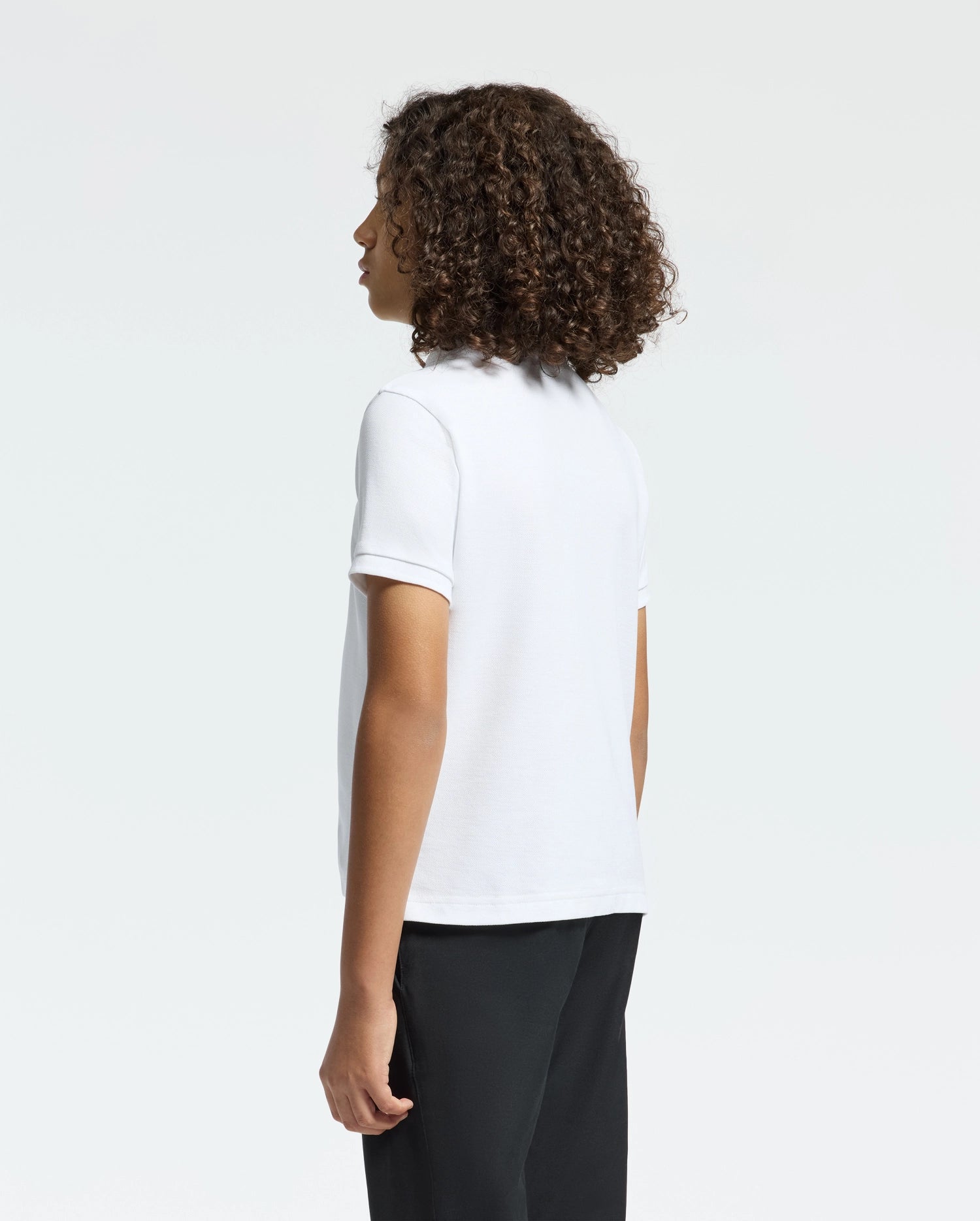 A person with curly hair, seen from the side and facing away, wears a white Psycho Bunny Kids Classic Pique Polo Shirt (B0K001CRPC) paired with black pants against a plain background.