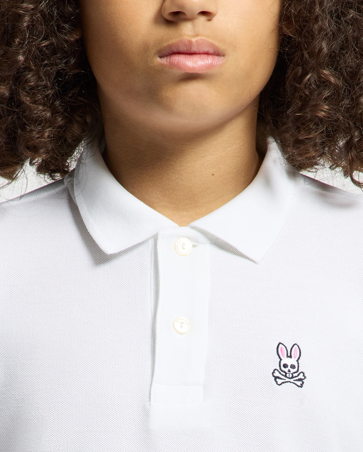 A person with curly hair is wearing a Psycho Bunny KIDS CLASSIC PIQUE POLO SHIRT in soft Pima cotton, adorned with a small pink bunny logo and crossbones on the left side. The image shows their upper torso and face, excluding the eyes.