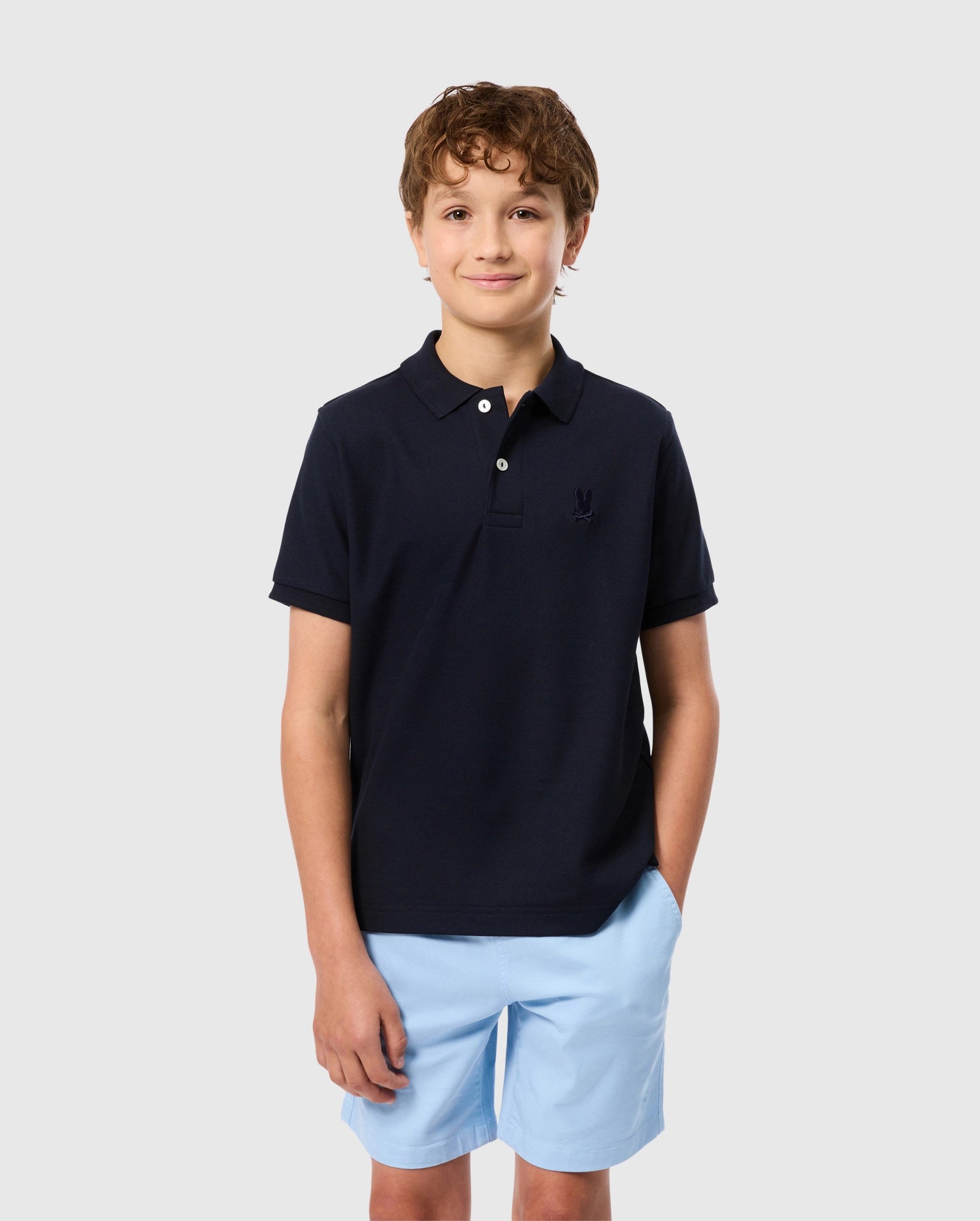 A young boy with curly hair is standing with his hands in the pockets of his light blue shorts. He is wearing a Psycho Bunny Kids Classic Pique Polo Shirt in dark navy, featuring mother-of-pearl buttons, and he is smiling slightly. The background is plain light gray.