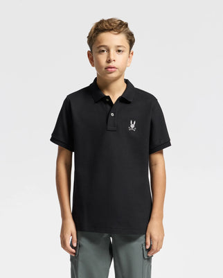 A young boy stands against a white background, wearing a black Psycho Bunny KIDS ALEXANDER PIQUE POLO (B0K101E200) with a small bunny logo on the chest. His light brown hair matches his neutral expression and relaxed posture, embodying effortless style.