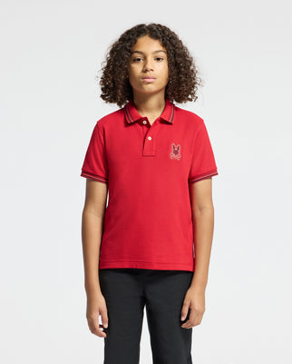 A young person with curly hair is wearing a red KIDS PIERCE PIQUE POLO - B0K201E200 from Psycho Bunny, adorned with an embroidered bunny on the chest, standing against a plain white background. Paired with black pants, this stylish ensemble highlights the playful charm of youthful fashion.