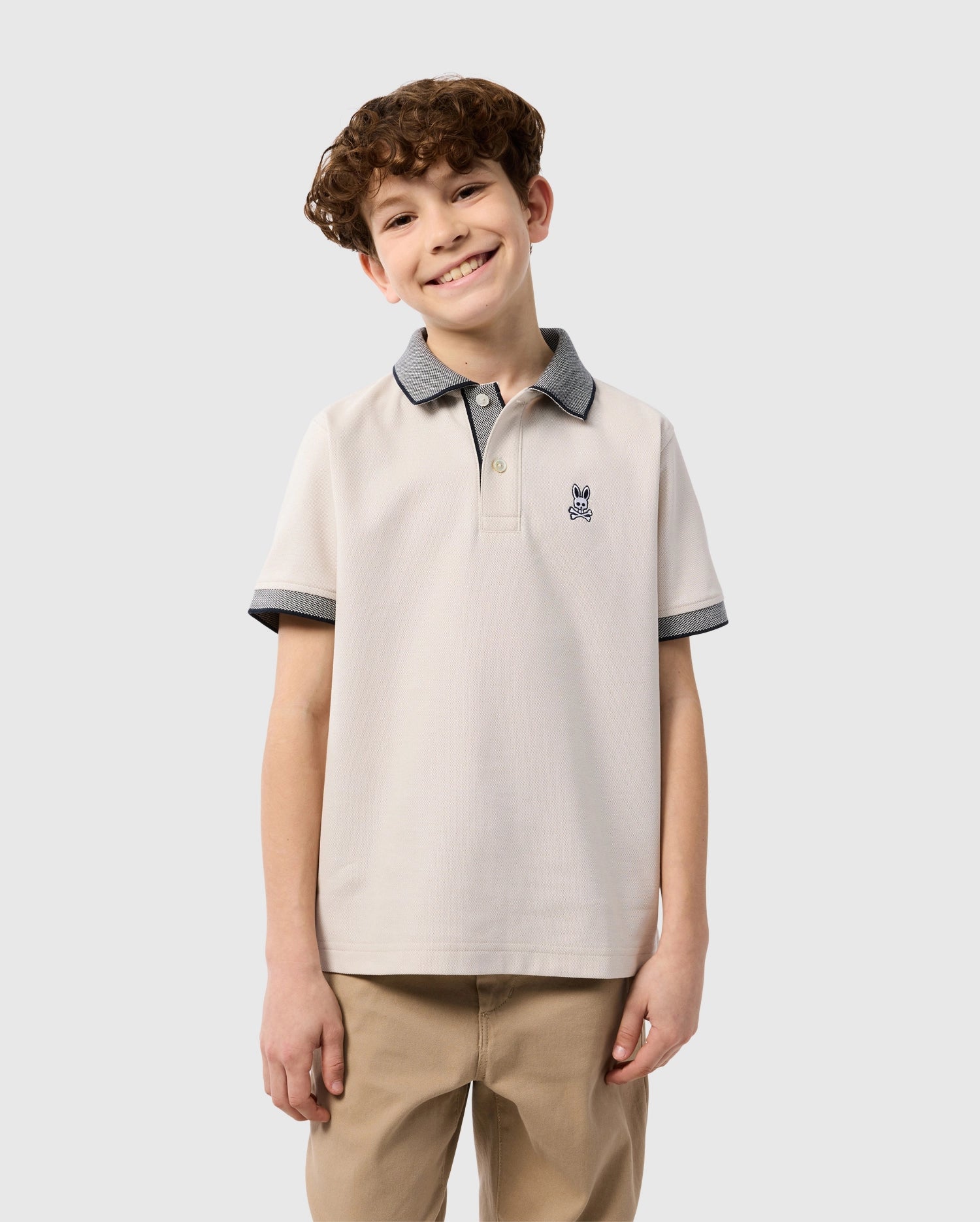 A smiling young boy with curly hair stands against a plain background, wearing a Psycho Bunny Kids Southport Pique Polo Shirt (B0K263B200) in light beige, crafted from Pima cotton piqué with a small embroidered logo and gray trim on the collar and sleeves, paired with khaki pants.