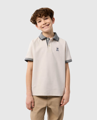 A smiling young boy with curly hair stands against a plain background, wearing a Psycho Bunny Kids Southport Pique Polo Shirt (B0K263B200) in light beige, crafted from Pima cotton piqué with a small embroidered logo and gray trim on the collar and sleeves, paired with khaki pants.