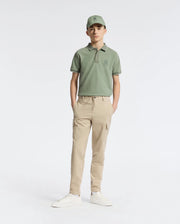 A teen boy wears a Psycho Bunny green cap and polo shirt, KIDS GREEN VALLEY CARGO PANT - B0P199E200 with ample pockets, and white sneakers while standing comfortably against a plain white background with hands in pockets.