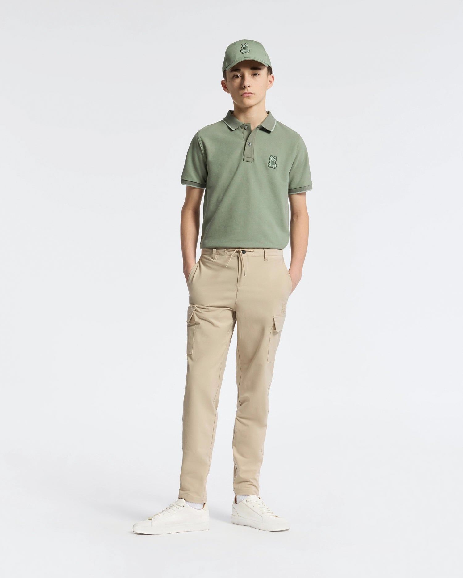 A teen boy wears a Psycho Bunny green cap and polo shirt, KIDS GREEN VALLEY CARGO PANT - B0P199E200 with ample pockets, and white sneakers while standing comfortably against a plain white background with hands in pockets.