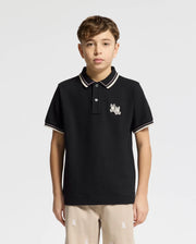 A boy is wearing the Psycho Bunny KIDS ELLIOT PIQUE POLO (B0K505F200) in black, featuring a chest Bunny logo and striped collar and sleeve details, paired with beige pants. He has short brown hair against a plain white background.