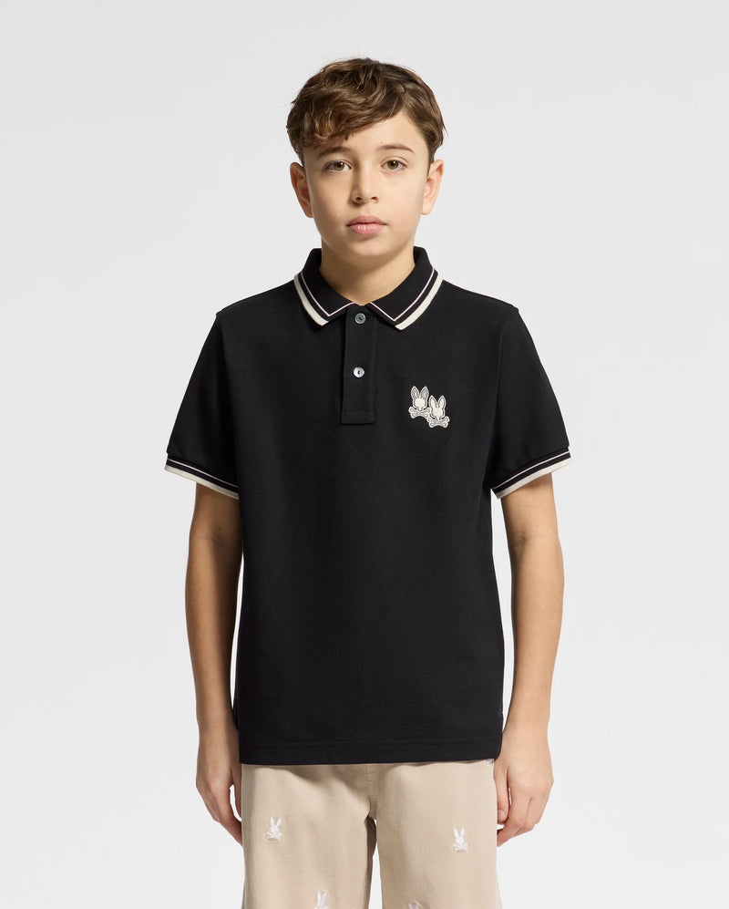 A boy is wearing the Psycho Bunny KIDS ELLIOT PIQUE POLO (B0K505F200) in black, featuring a chest Bunny logo and striped collar and sleeve details, paired with beige pants. He has short brown hair against a plain white background.