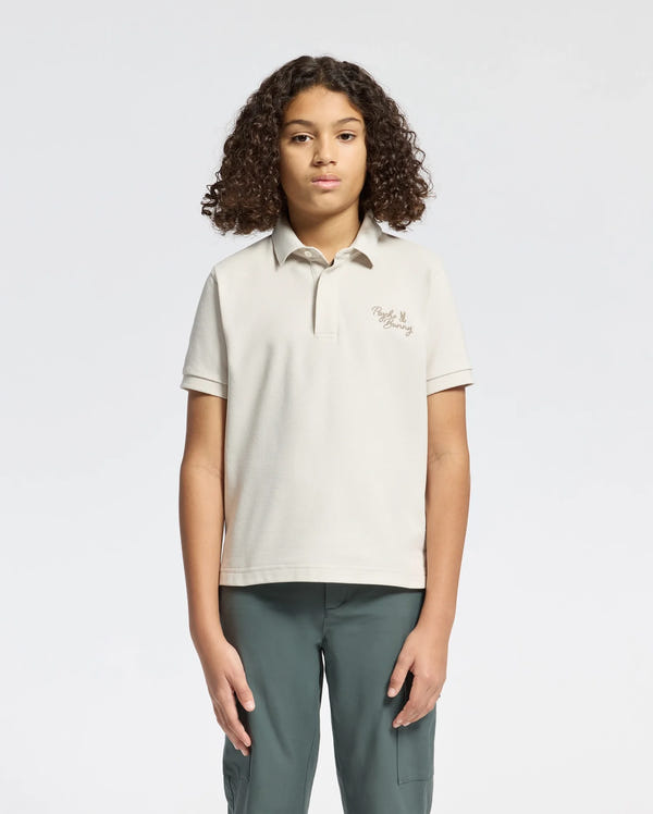 A child with curly hair wears a Psycho Bunny KIDS DESERT PIQUE POLO - B0K506F200, made of 100% Pima cotton, along with dark green pants against a plain white background. The beige polo features an embroidered design on the upper left, conveying timeless style.