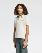 A person with curly hair faces forward, wearing Psycho Bunny's timeless KIDS DESERT PIQUE POLO - B0K506F200 made from 100% Pima cotton. The light beige shirt has a small logo on the left and is perfectly paired with dark green pants against a plain white background.
