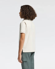 A person with curly hair is seen from the side wearing a Psycho Bunny KIDS DESERT PIQUE POLO made of 100% Pima cotton and green cargo pants, exuding timeless style against a plain, light-colored background.