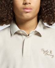 A person sporting the KIDS DESERT PIQUE POLO by Psycho Bunny with a brown embroidered logo on the chest. Their timeless style, enhanced by curly hair, is perfectly completed by 100% Pima cotton, though only the lower part of their face is visible.