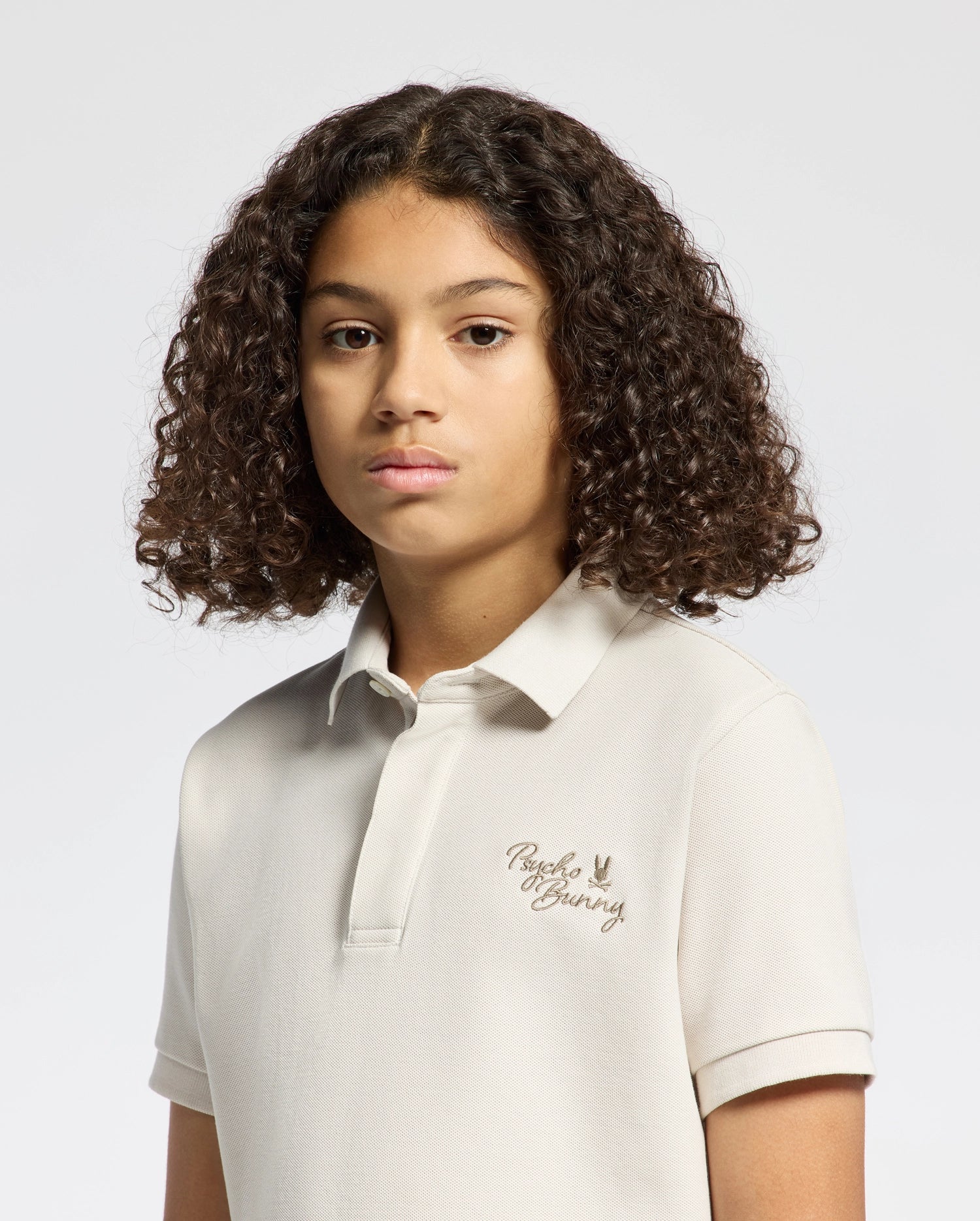 A young individual with curly brown hair wears the Psycho Bunny KIDS DESERT PIQUE POLO in light beige, crafted from 100% Pima cotton and adorned with a small logo on the left. The classic look is accented against a simple, light background.