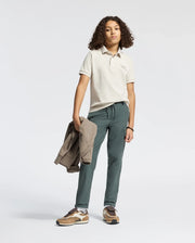 A curly-haired teen confidently sports Psycho Bunny's KIDS DESERT PIQUE POLO – B0K506F200, made from 100% Pima cotton. Paired with dark pants and a jacket in one hand, brown sneakers finish the casual look against a plain white backdrop.
