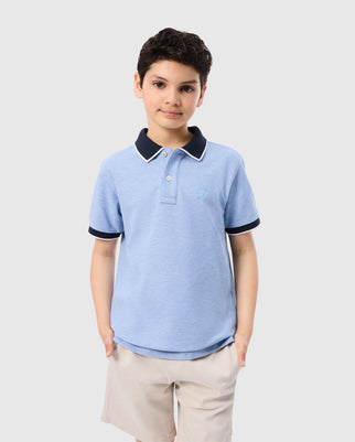 A young boy stands against a plain background wearing a light blue KIDS WINDCREST PIQUE POLO SHIRT - B0K592C200 by Psycho Bunny with a navy collar and sleeve cuffs, paired with beige shorts. He has short, dark curly hair and his hands are tucked into his shorts pockets. The embroidered chest Bunny adds a delightful touch to his high-quality outfit.