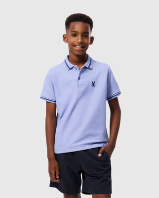 A young boy smiles, standing with one hand in his pocket. He wears a light purple Psycho Bunny KIDS RIVIERA PIQUE POLO SHIRT - B0K640C200 made from diamond-knit Pima cotton, featuring an embroidered chest Bunny and dark blue stripes on the collar and sleeves. He pairs the shirt with dark blue shorts. The background is plain and light-colored.