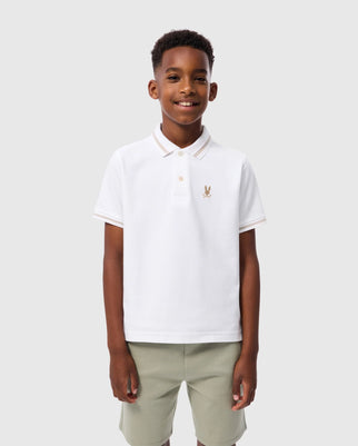A boy stands against a plain, light grey background, smiling. He is wearing a white Psycho Bunny KIDS RIVIERA PIQUE POLO SHIRT - B0K640C200 with an embroidered chest Bunny logo and light green shorts. His hair is short and neatly styled.