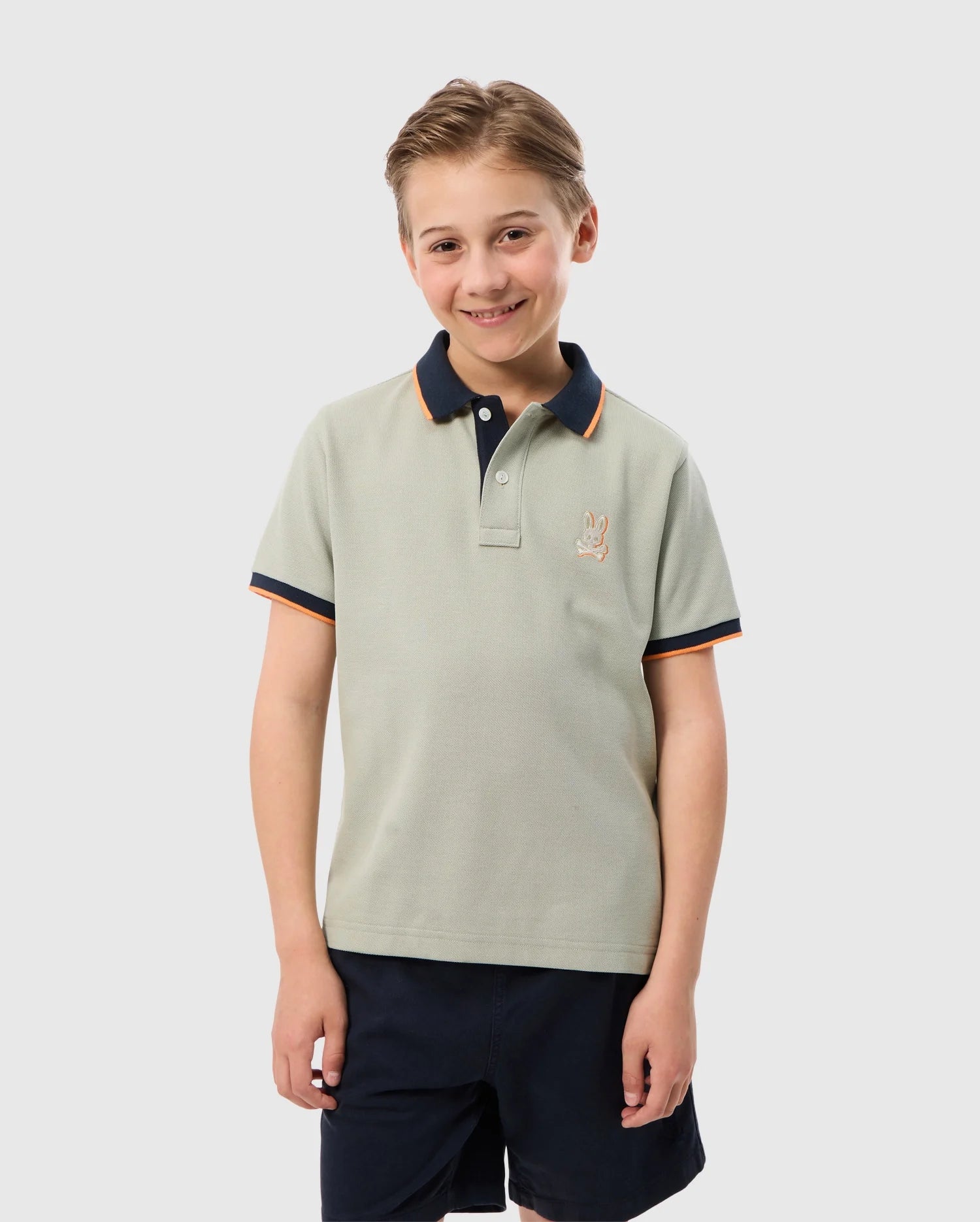 A young boy with short blond hair is smiling and standing with his arms at his sides. He is wearing a light green KIDS KAYDEN PIQUE POLO - B0K675C200 by Psycho Bunny, which features a dark blue collar and rib knit cuffs. His shorts complement the outfit, while a small embroidered rabbit logo decorates the left chest of the polo shirt. The background is plain white.