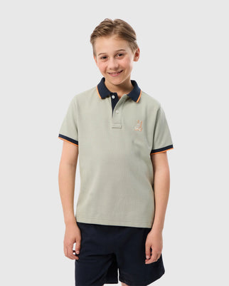 A young boy with short blond hair is smiling and standing with his arms at his sides. He is wearing a light green KIDS KAYDEN PIQUE POLO - B0K675C200 by Psycho Bunny, which features a dark blue collar and rib knit cuffs. His shorts complement the outfit, while a small embroidered rabbit logo decorates the left chest of the polo shirt. The background is plain white.