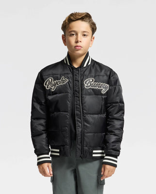 A young boy stands wearing the stylish KIDS FRASER FASHION PUFFER - B0N338E200 from Psycho Bunny, a black puffer jacket with embroidered "Psycho Bunny" text on both sides of the chest. This fashionable kids' puffer showcases striped cuffs and collar. He has short hair and maintains a neutral expression against a plain background.