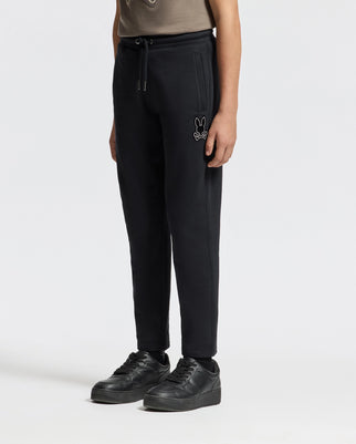 A person wearing Psycho Bunny's KIDS ALEXANDER SWEATPANT in black, featuring their signature bunny logo on the left side, paired with a dark gray top and black sneakers, stands against a plain white background.