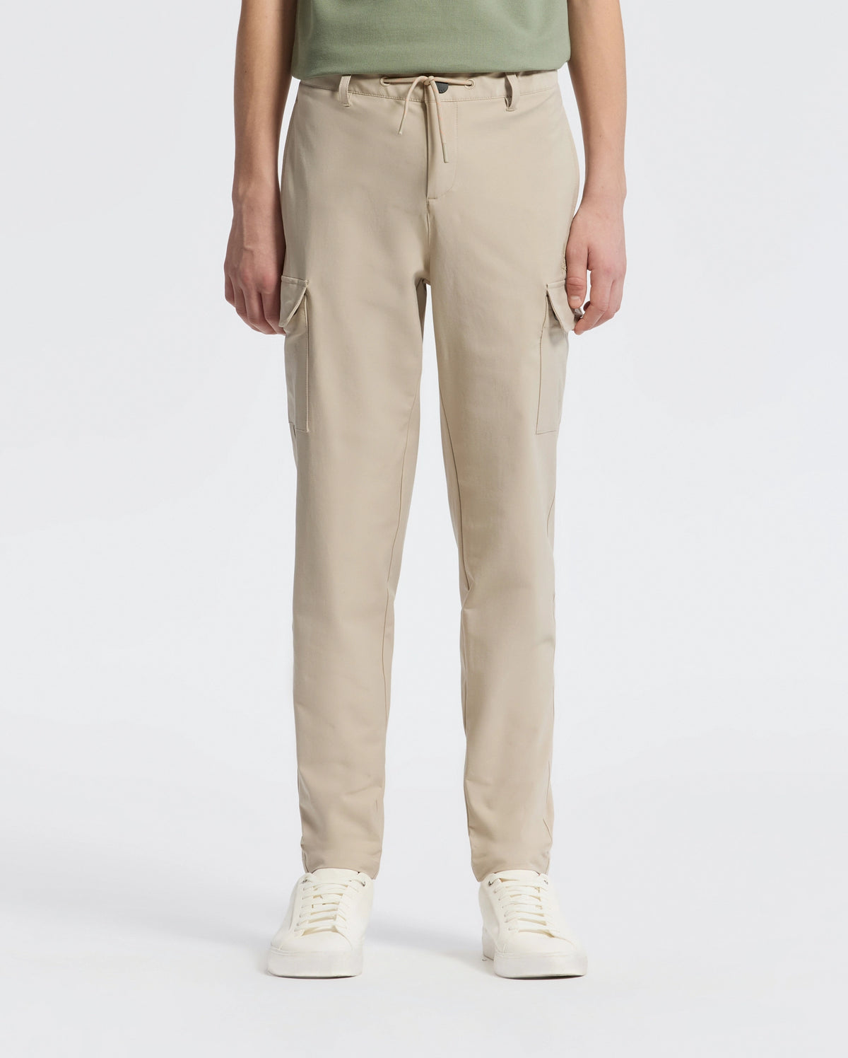 A person stands against a plain backdrop wearing Psycho Bunny’s KIDS GREEN VALLEY CARGO PANT - B0P199E200, featuring beige drawstring waist and multiple pockets, paired with white sneakers. The upper body is partially visible.