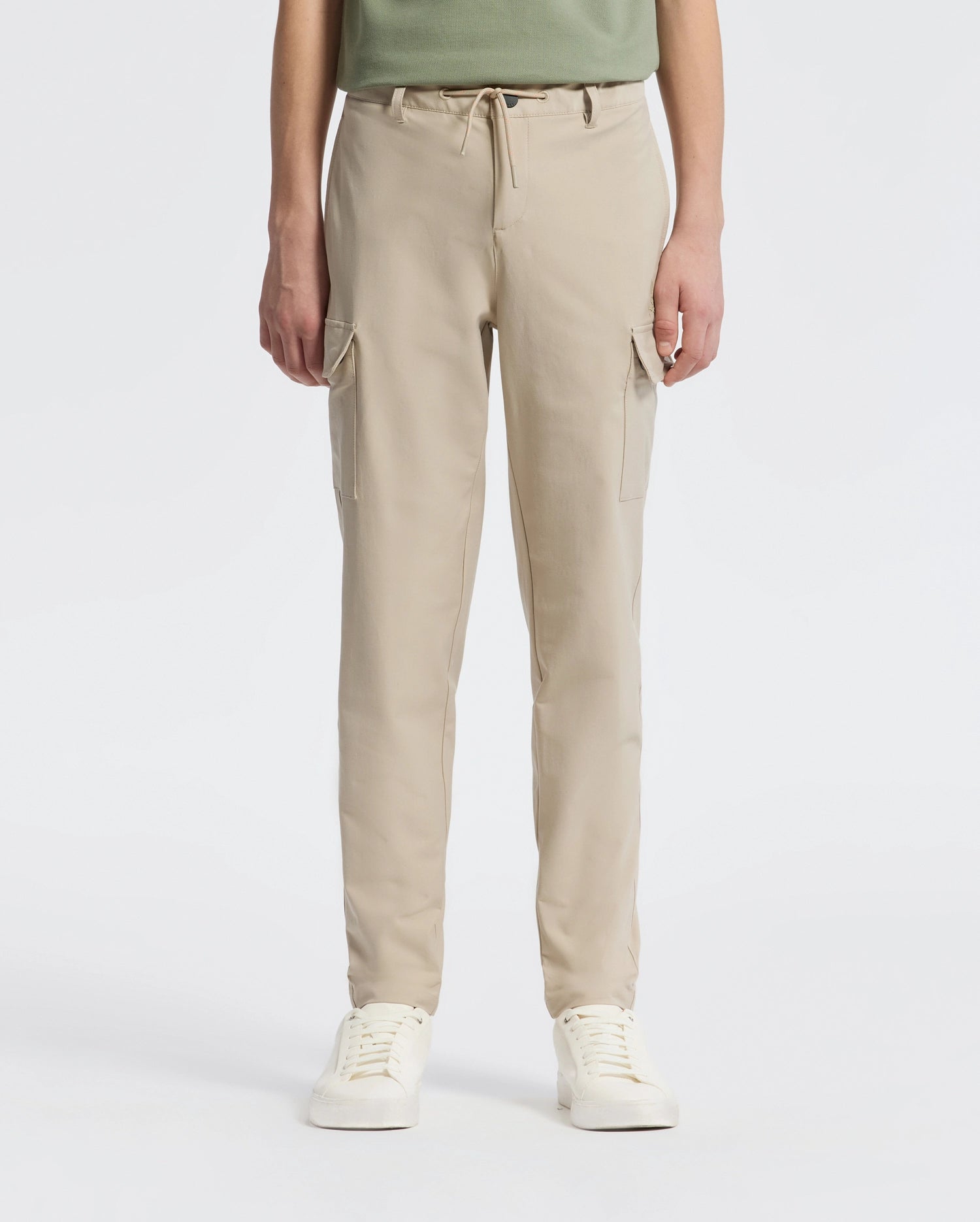 A person stands against a plain backdrop wearing Psycho Bunny’s KIDS GREEN VALLEY CARGO PANT - B0P199E200, featuring beige drawstring waist and multiple pockets, paired with white sneakers. The upper body is partially visible.
