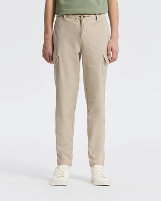 A person stands against a plain backdrop wearing Psycho Bunny’s KIDS GREEN VALLEY CARGO PANT - B0P199E200, featuring beige drawstring waist and multiple pockets, paired with white sneakers. The upper body is partially visible.