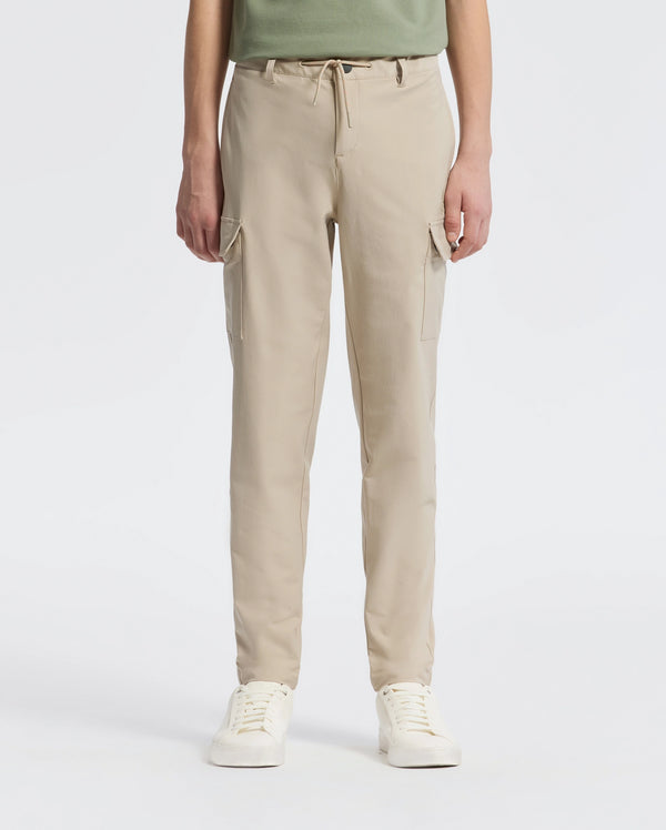 A person stands against a plain backdrop wearing Psycho Bunny’s KIDS GREEN VALLEY CARGO PANT - B0P199E200, featuring beige drawstring waist and multiple pockets, paired with white sneakers. The upper body is partially visible.