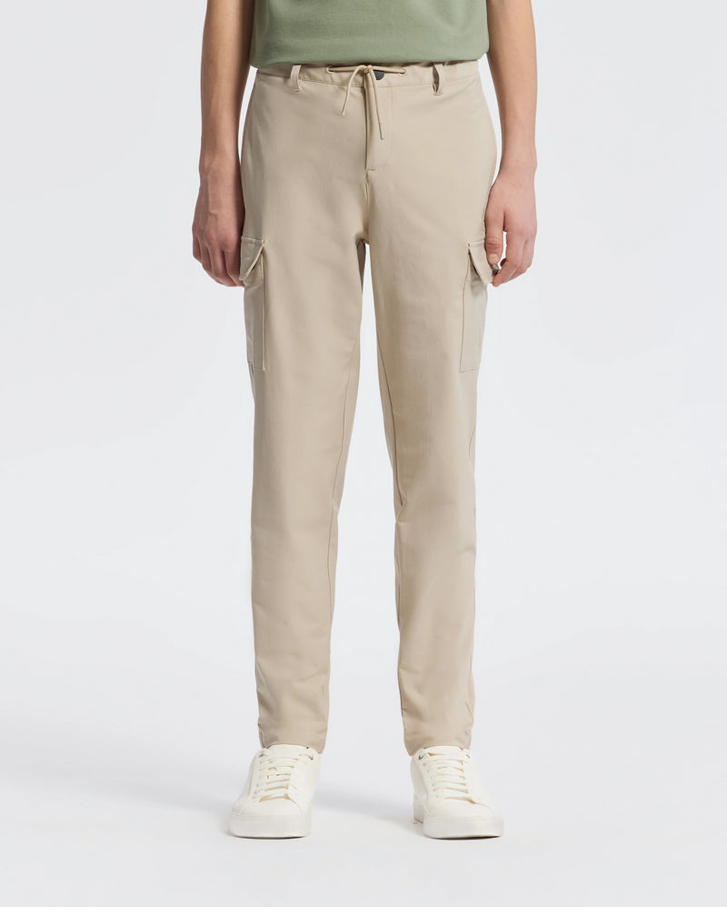 A person stands against a plain backdrop wearing Psycho Bunny’s KIDS GREEN VALLEY CARGO PANT - B0P199E200, featuring beige drawstring waist and multiple pockets, paired with white sneakers. The upper body is partially visible.