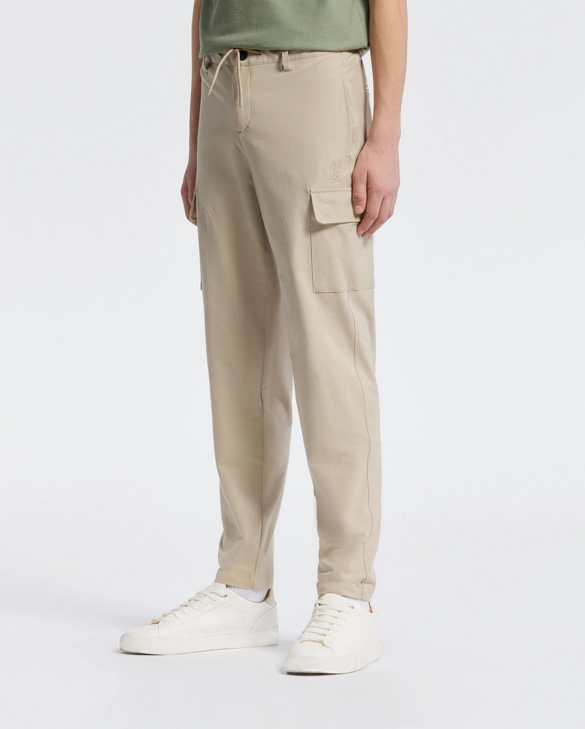 Against a white background, a person models the chic and practical KIDS GREEN VALLEY CARGO PANT by Psycho Bunny. The beige cargo pants feature multiple pockets, perfectly styled with white sneakers and a green shirt for an effortlessly stylish look, highlighting their practicality.