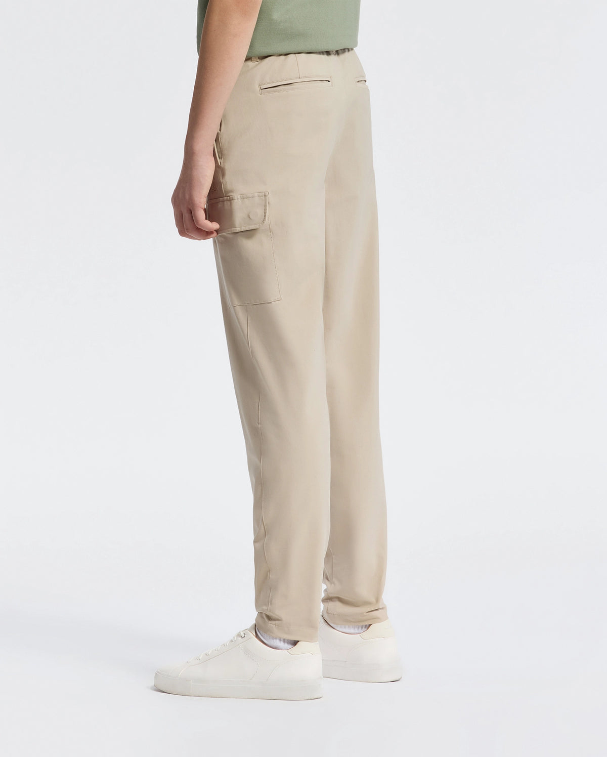 A person wearing Psycho Bunny's Kids Green Valley Cargo Pant (B0P199E200) with side pockets, paired with white sneakers, stands sideways against a plain white background. They're dressed in a green top that's partially visible.