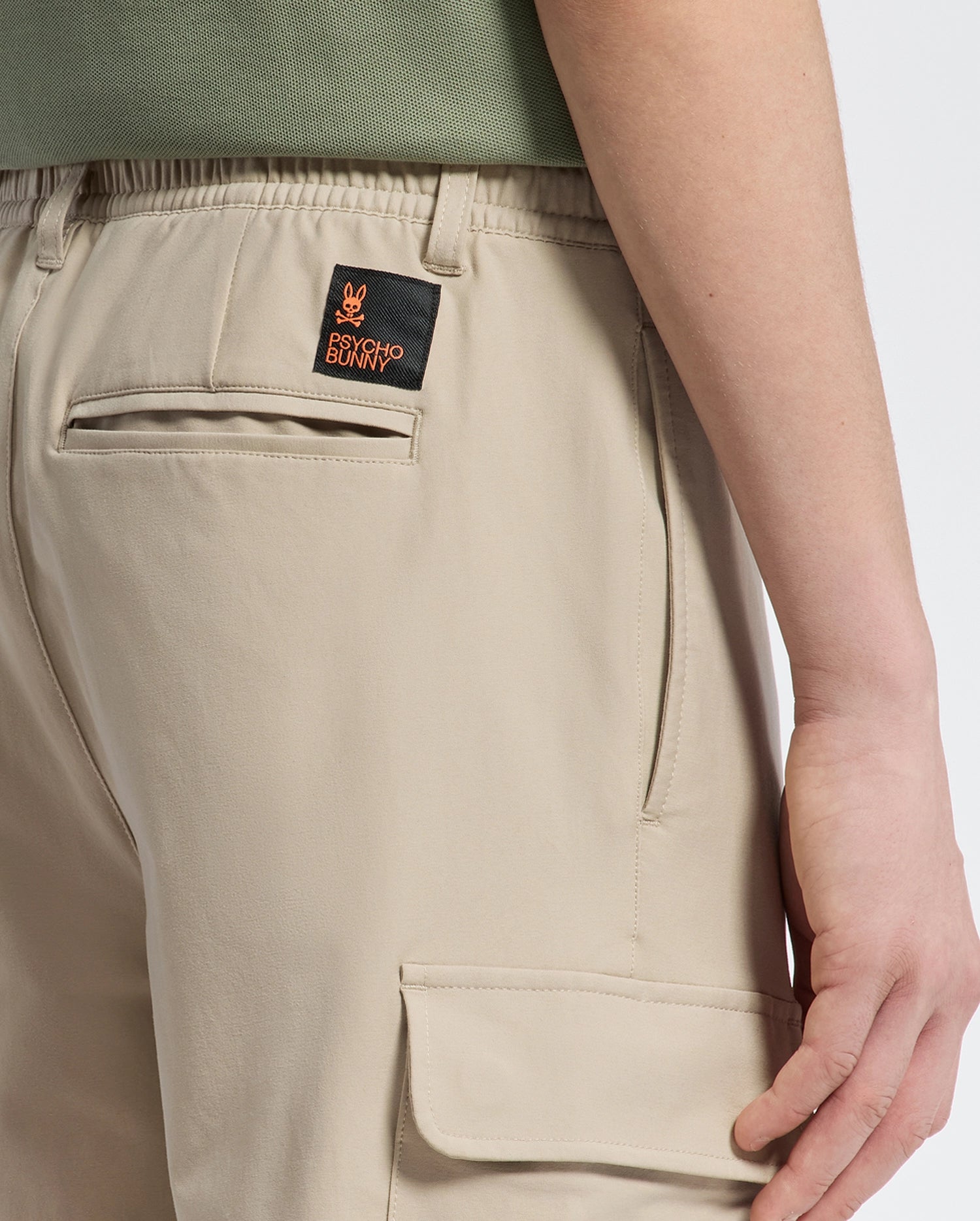 A person is shown from the side in Psycho Bunny's KIDS GREEN VALLEY CARGO PANT (B0P199E200) featuring beige fabric, practical pockets, and an embroidered patch with a small orange design on the back. Paired with a green shirt, this look combines style and comfort seamlessly.