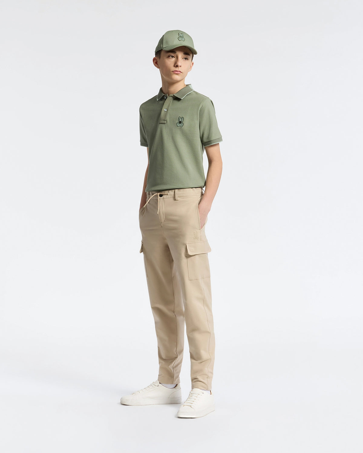 A person wearing a green polo shirt, matching cap, Psycho Bunny KIDS GREEN VALLEY CARGO PANT - B0P199E200 with ample pockets, and white sneakers stands against a plain white background. One hand is in their pocket as they appear comfortably relaxed.