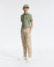 A person wearing a green polo shirt, matching cap, Psycho Bunny KIDS GREEN VALLEY CARGO PANT - B0P199E200 with ample pockets, and white sneakers stands against a plain white background. One hand is in their pocket as they appear comfortably relaxed.