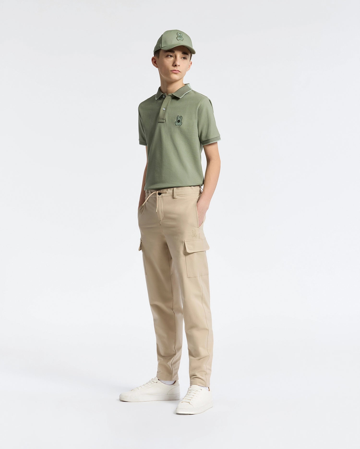 A person wearing a green polo shirt, matching cap, Psycho Bunny KIDS GREEN VALLEY CARGO PANT - B0P199E200 with ample pockets, and white sneakers stands against a plain white background. One hand is in their pocket as they appear comfortably relaxed.