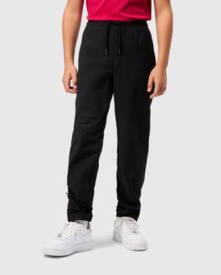 A person in a red shirt, black Psycho Bunny Kids Madison Commuter Pants (B0P395Z1WB), and white sneakers stands against a plain background. The joggers have an elastic waistband with a drawstring, elastic cuffs at the ankles, and hidden Bunny detailing for a playful touch.