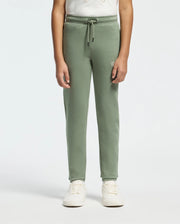 A child wearing Psycho Bunny's KIDS DESERT EMBROIDERED MODAL SWEATPANT (B0P489F200) in light green, featuring an embroidered wordmark and a drawstring, is paired with a light t-shirt and white sneakers. The background is plain and neutral.