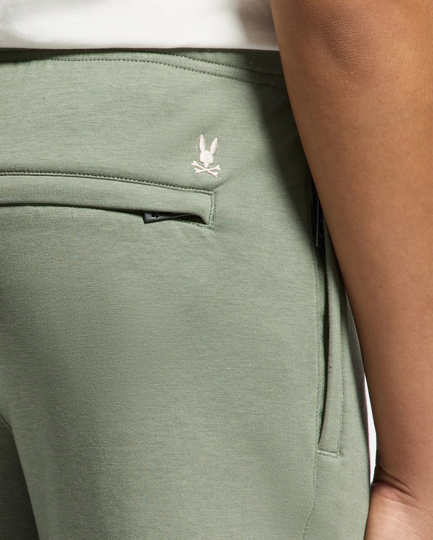 A close-up displays the olive green KIDS DESERT EMBROIDERED MODAL SWEATPANT - B0P489F200 by Psycho Bunny, featuring a visible pocket and embroidered bunny logo above crossed bones near the waistband. Crafted from soft stretch jersey, they show a glimpse of a white shirt at the waist.