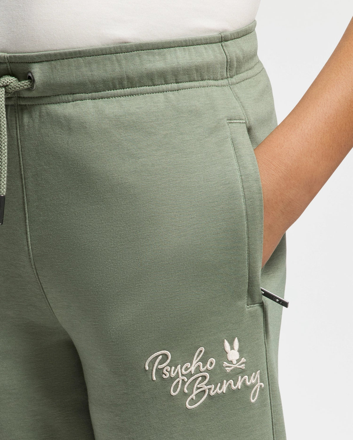 A person is wearing modal-blend sweatpants from Psycho Bunny, featuring the "Desert" embroidery near the pocket. These KIDS DESERT EMBROIDERED MODAL SWEATPANT (B0P489F200) are crafted from soft stretch jersey with an elastic waistband and drawstring, and they have a hand resting in the pocket.