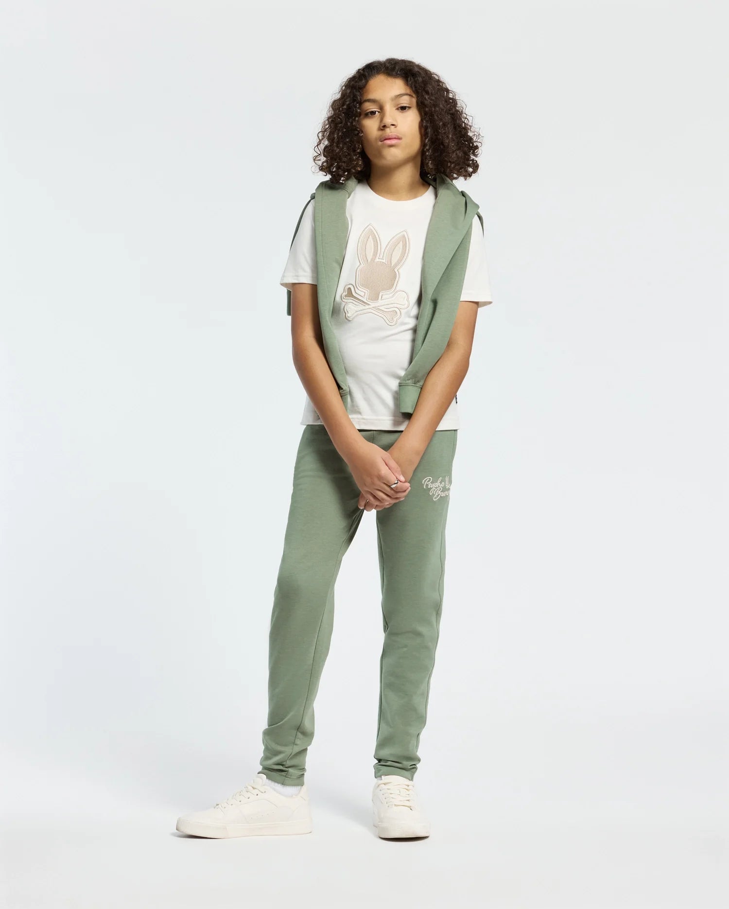 A teen is wearing a white T-shirt with an embroidered wordmark, a green sleeveless hoodie, and Psycho Bunny's KIDS DESERT EMBROIDERED MODAL SWEATPANT in green. The outfit features modal-blend sweatpants and soft stretch jersey material. Completing the look are white sneakers against a plain backdrop.