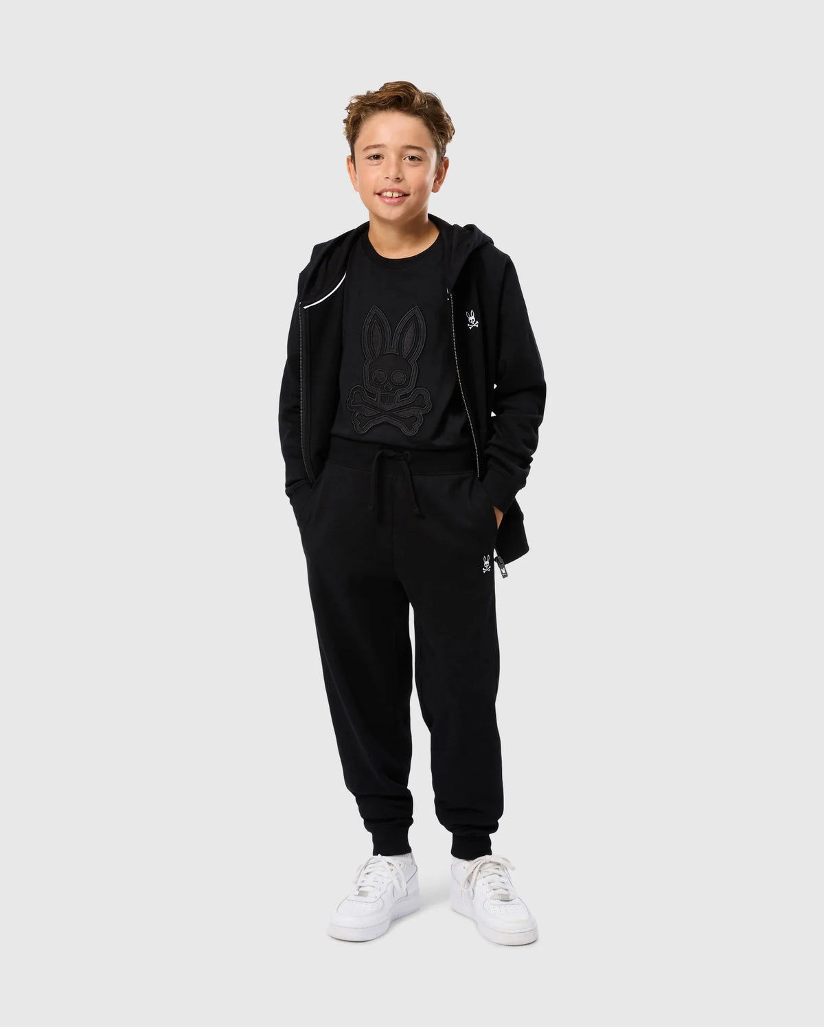 A young person in a black hoodie and Kids Levy Essential Fleece Sweatpants by Psycho Bunny, featuring a cartoon bunny design, stands smiling in white sneakers against a plain white background.