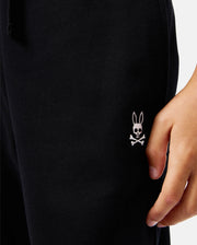 Dressed in Psycho Bunny KIDS LEVY ESSENTIAL FLEECE SWEATPANTS, black with a white rabbit skull and crossbones logo by the pocket, this person exudes style and comfort as their hand rests casually on the side.