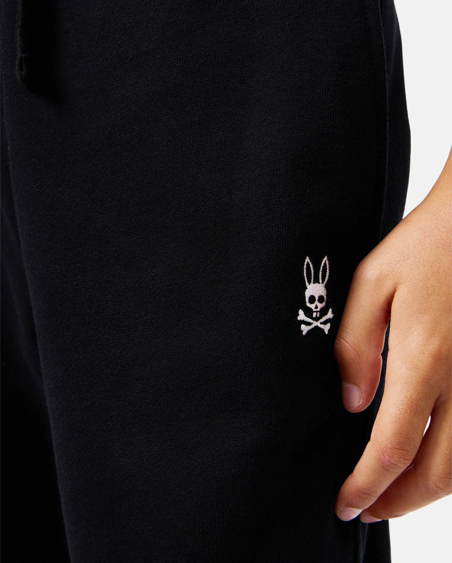 Dressed in Psycho Bunny KIDS LEVY ESSENTIAL FLEECE SWEATPANTS, black with a white rabbit skull and crossbones logo by the pocket, this person exudes style and comfort as their hand rests casually on the side.